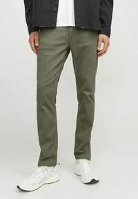 Pantaloni in cotone verde oliva con vestibilità dritta e tasche frontali, indossati con scarpe da ginnastica bianche. Tessuto liscio e design semplice.