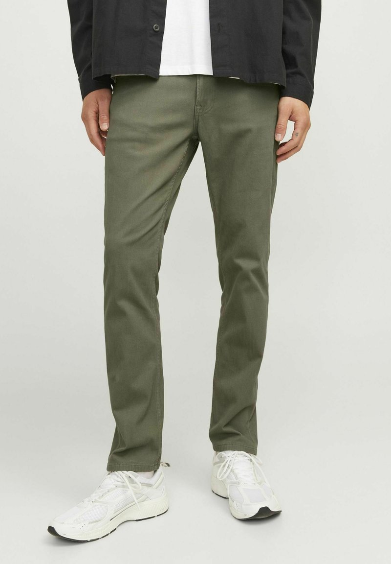 Pantaloni in cotone verde oliva con vestibilità dritta e tasche frontali, indossati con scarpe da ginnastica bianche. Tessuto liscio e design semplice.