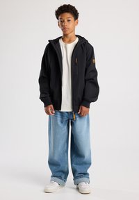 Schwarze Softshelljacke mit Kapuze, Reißverschluss-Tasche und orangefarbenem Reißverschluss-Zipper, kombiniert mit lockeren blauen Jeans und weißen Sneakern.