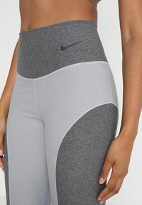 Szare legginsy z wysokim stanem z czarnym logo Nike. Posiadają wzór w blokach kolorystycznych, gładką teksturę oraz kontrastowe wstawki.