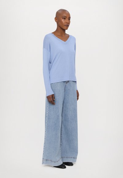 Maglione azzurro chiaro a V in un morbido filato, abbinato a jeans in denim chiaro a gamba larga. L'outfit presenta una silhouette rilassata e delicati dettagli di cucitura.