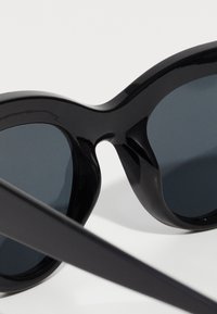 Mustat muoviset aurinkolasit, joissa on pyöristetty kissansilmämuoto. Tummat linssit ja sileä pinta, jossa on paksu ja kaareva kehysdesign.