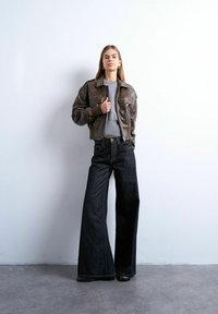 Jeune femme debout contre un mur blanc, portant une veste en cuir marron, un pull gris et un jean noir à jambes larges avec des chaussures noires.