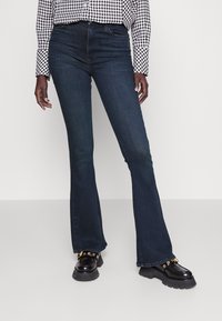 Persona vestida con jeans de campana azul oscuro, mocasines negros gruesos con detalles de ojales dorados, y una blusa de cuadros en blanco y negro, de pie sobre un suelo blanco.