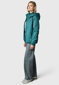 Vrouw met een turquoise hoodie, grijze jeans met wijde pijpen en grijze sneakers, zijwaarts staand tegen een effen lichte achtergrond.