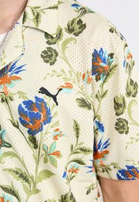 Blomster kortærmet skjorte i lysebeige mesh-stof, med blå, orange og grønne mønstre, samt et sort Puma-logo på brystet.