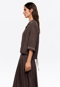 Brązowa krótka koszula z krótkimi rękawami i luźnym krojem, posiadająca teksturowane wykończenie oraz subtelne przeszycia na mankietach.