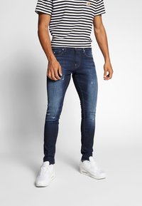 Dunkelblaue Jeans im Slim-Fit, mit Destroyed-Details und glatter Textur. Kombiniert mit einem schwarz-weißen gestreiften T-Shirt.