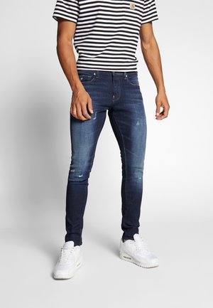 Mörkblå jeans med slim fit, med slitna detaljer och en mjuk textur. Matchas med en svart- och vitrandig t-shirt.