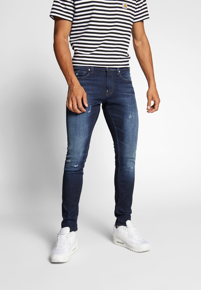 Dunkelblaue Jeans im Slim-Fit, mit Destroyed-Details und glatter Textur. Kombiniert mit einem schwarz-weißen gestreiften T-Shirt.