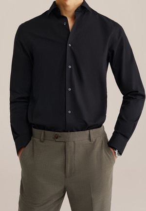 Hombre vistiendo una camisa negra de manga larga abotonada, metida dentro de pantalones marrones a medida, con las manos en los bolsillos y un reloj de pulsera plateado.