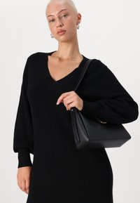 Svart stickad klänning med V-ringning och lösa ärmar. Modellen håller en svart strukturerad handväska med ett guldaccent. Slät textur, minimal design.