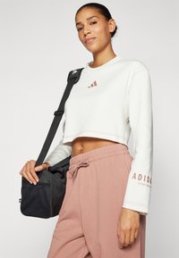 Balta trumpa sportinė palaidinė su ruda Adidas logotipu, derinama su rožinėmis treningo kelnėmis. Modelis laiko juodą pečių krepšį su tekstūruota paviršiumi.