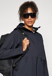 Veste noire imperméable avec fermeture éclair à l'avant, capuche ajustable et plusieurs poches, portée avec des lunettes de soleil surdimensionnées et un sac à dos.