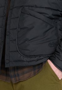 Chaqueta de plumas negra con textura cosida y cierre de botón, que cuenta con un gran bolsillo y un logo de árbol bordado en el lateral.
