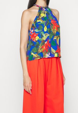 Frau trägt ärmelloses Neckholder-Top mit hellem Blumenmuster in Blau, Rot, Gelb und Grün, kombiniert mit locker sitzender, leuchtend roter Hose.