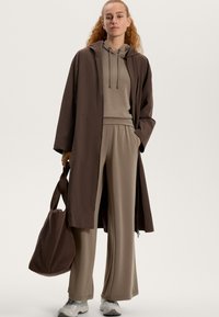 Brązowy lekki płaszcz na beżowym hoodie oraz dopasowanych szerokich spodniach. Zawiera brązową torbę na ramie i szare sneakersy. Gładka faktura materiału.