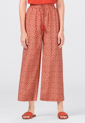 Femme portant un pantalon large rouge à motif floral avec taille à cordon et des sandales rouges ouvertes à l'avant sur fond uni.