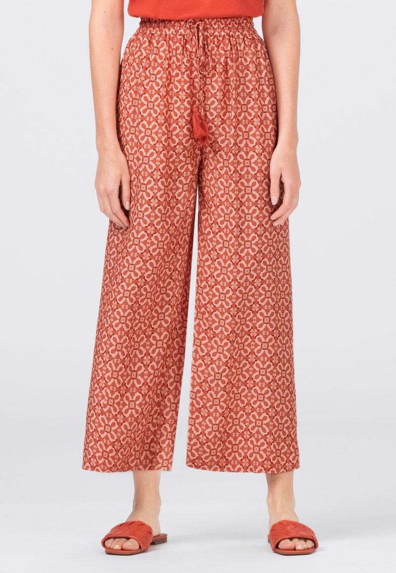 Femme portant un pantalon large rouge à motif floral avec taille à cordon et des sandales rouges ouvertes à l'avant sur fond uni.