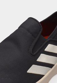 Μαύρο αθλητικό παπούτσι χωρίς κορδόνια με υφασμάτινη επιφάνεια με υφή, λευκές λοξές ρίγες στο πλευρό και μπεζ σόλα που διακρίνεται πάνω σε ανοιχτό φόντο.