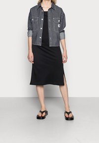 Jeansjacka med vita ränder, knäppa, och bröstfickor över en svart, ärmlös klänning med en sidoöppning, ihop med svarta sandaler.