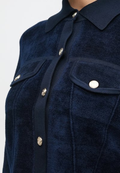 Chaqueta texturizada color azul marino con cuello, cierre de botón en el frontal y dos bolsillos en el pecho con botones dorados brillantes.