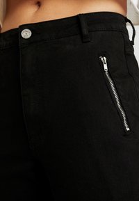 Pantalones de mezclilla negra con un bolsillo frontal con cremallera, que cuenta con un botón plateado y costuras elegantes. La tela tiene una textura suave.