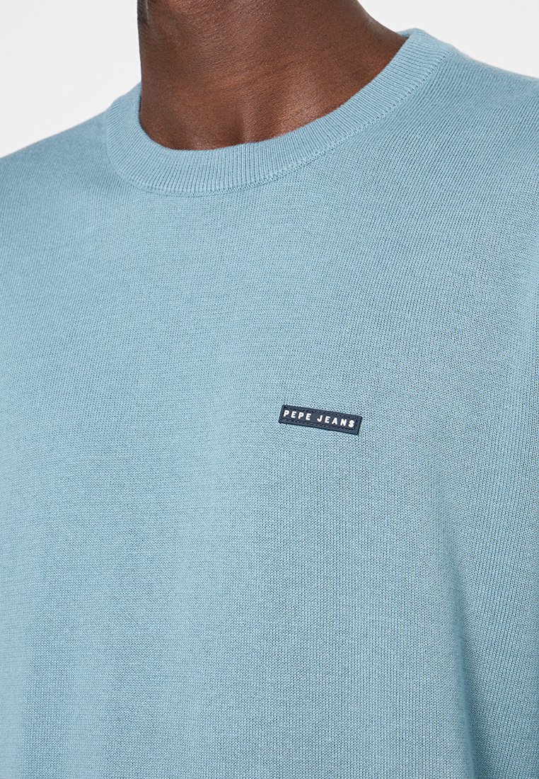 Jasnoniebieski sweter z okrągłym dekoltem, wykonany z dzianiny, z małą czarną metką z napisem "PEPE JEANS" na klatce piersiowej. Gładka tekstura, prosty design.