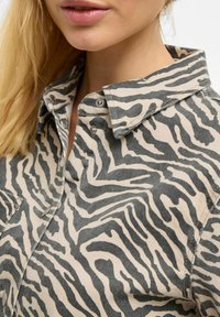 Knappskjorte i beige stoff med et dristig svart zebra-stripe mønster, med klassisk krage og sølvfargede trykknapper.