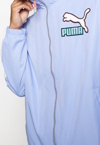 Chaqueta azul claro con tejido texturizado, que presenta una cremallera plateada, logo de Puma bordado en blanco y turquesa, y puños elásticos.