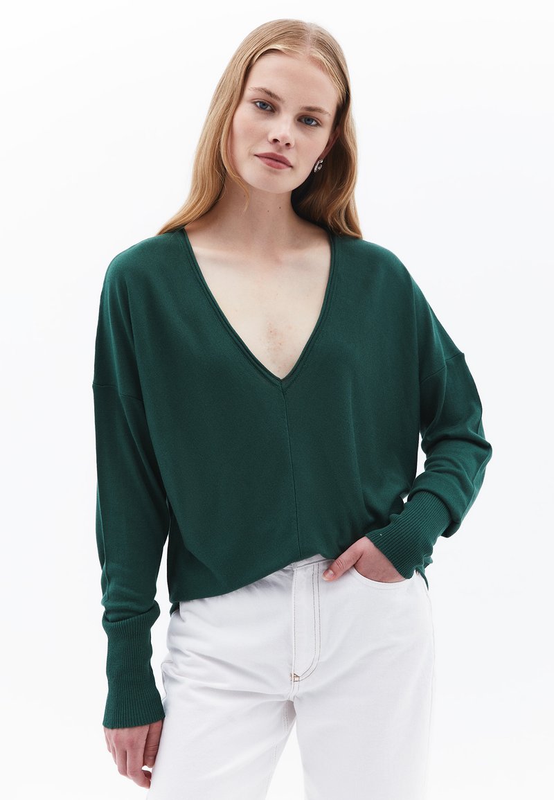 Grüner, weicher Stoffpullover mit tiefem V-Ausschnitt, langen Ärmeln und gerippten Bündchen, kombiniert mit weißen Hosen. Einfaches Design ohne Muster.