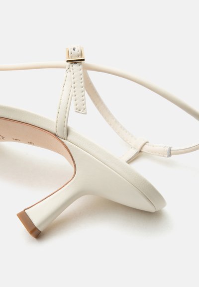 Witte leren sandaal met hoge hak, dunne enkelband en kleine gouden gesp op een effen witte achtergrond.