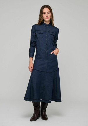Donna che indossa un vestito lungo di denim blu scuro con bottoni frontali e maniche lunghe, abbinato a stivali marrone scuro, in piedi su uno sfondo semplice.