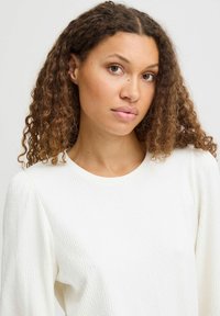 Cremefarbener, strukturierten Pullover mit Rippmuster, Rundhalsausschnitt und leicht gepufften Ärmeln, getragen von einer Person mit lockigem Haar.