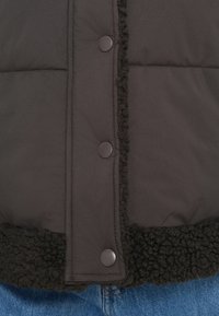 Melnā polsterētā veste ar tekstiturētu flīsa apakšmalu, ar noapaļotiem melniem pogām un izšuvumiem gar priekšējo paneli.