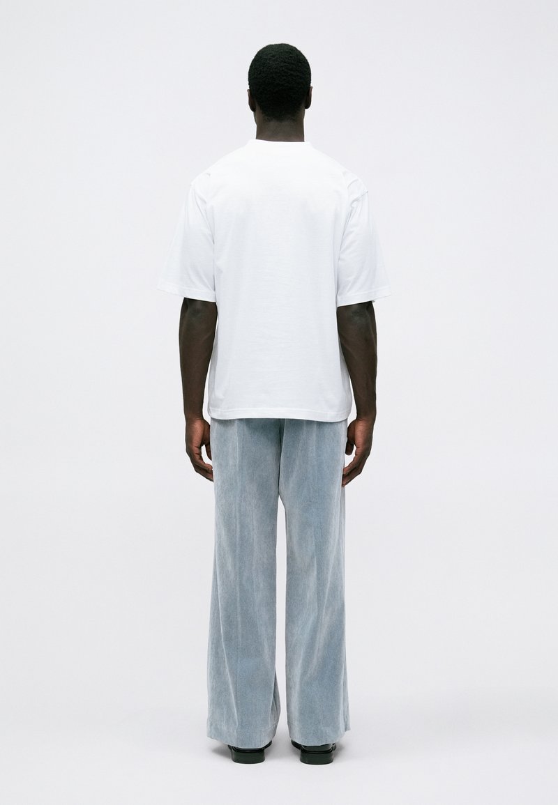 Homme se tenant de dos, portant un t-shirt blanc à manches courtes, un pantalon large gris clair et des chaussures noires sur un fond blanc.