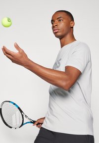 Grå treningst-skjorte med korte ermer, holder en tennisracket i den ene hånden mens han kaster en neongrønn tennisball.