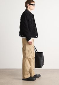 Black corduroy jacket, beige cargo pants, black footwear, and a black tote bag. Model wears sunglasses and stands against a neutral background.