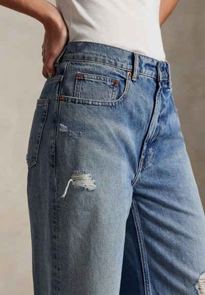 Lyseblå dongeribukser med høyt liv, slitte detaljer på låret og metallakenter på lommene. Teksturert stoff.