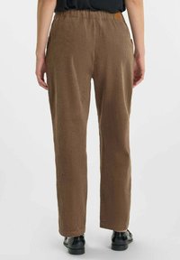 Persona que lleva pantalones de pana marrón de tiro alto y sueltos, y zapatos negros, visto desde atrás, contra un fondo liso.