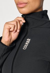 Giacca nera con zip realizzata in un tessuto liscio, dotata di un colletto alto e del logo "COLMAR" di colore chiaro sul davanti. Primo piano sulla zona del collo.