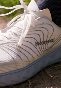 Weißer Sportschuh mit strukturierten Oberflächen, schwarzen Akzenten, dünnen weißen Schnürsenkeln und einer flexiblen Sohle mit gewelltem Muster.