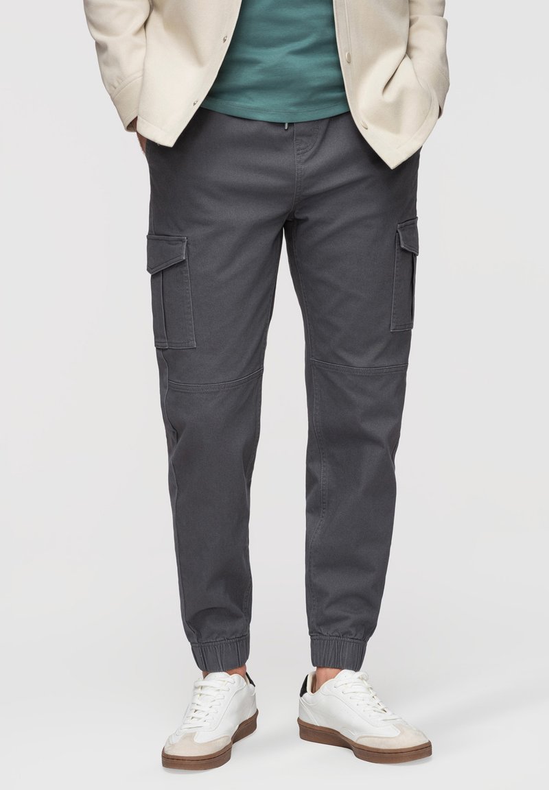 Homme portant un pantalon jogging cargo gris foncé avec des poignets élastiques, des baskets blanches, une chemise bleu sarcelle et une veste beige, les mains dans les poches.
