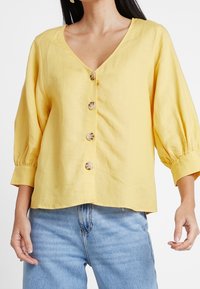 Geel knoop-overhemd met een V-hals, 3/4 mouwen en een losse pasvorm. Opvallende kenmerken zijn grote ronde knopen en een zachte textuur.