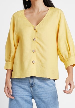 Haut boutonnière jaune avec un col en V, des manches 3/4 et une coupe ample. Les caractéristiques notables incluent de grands boutons ronds et une texture douce.