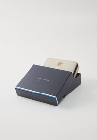 Cremefarbige Lederbörse mit goldenem Logo, präsentiert in einer navyblauen Box mit rot-weißen Streifenakzenten. Mit Reißverschluss und strukturiertem Oberflächen.