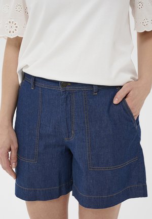 Witte top met korte mouwen en oogjesdetails, gecombineerd met blauwe denim shorts met grote voorzakken en zichtbare naden.