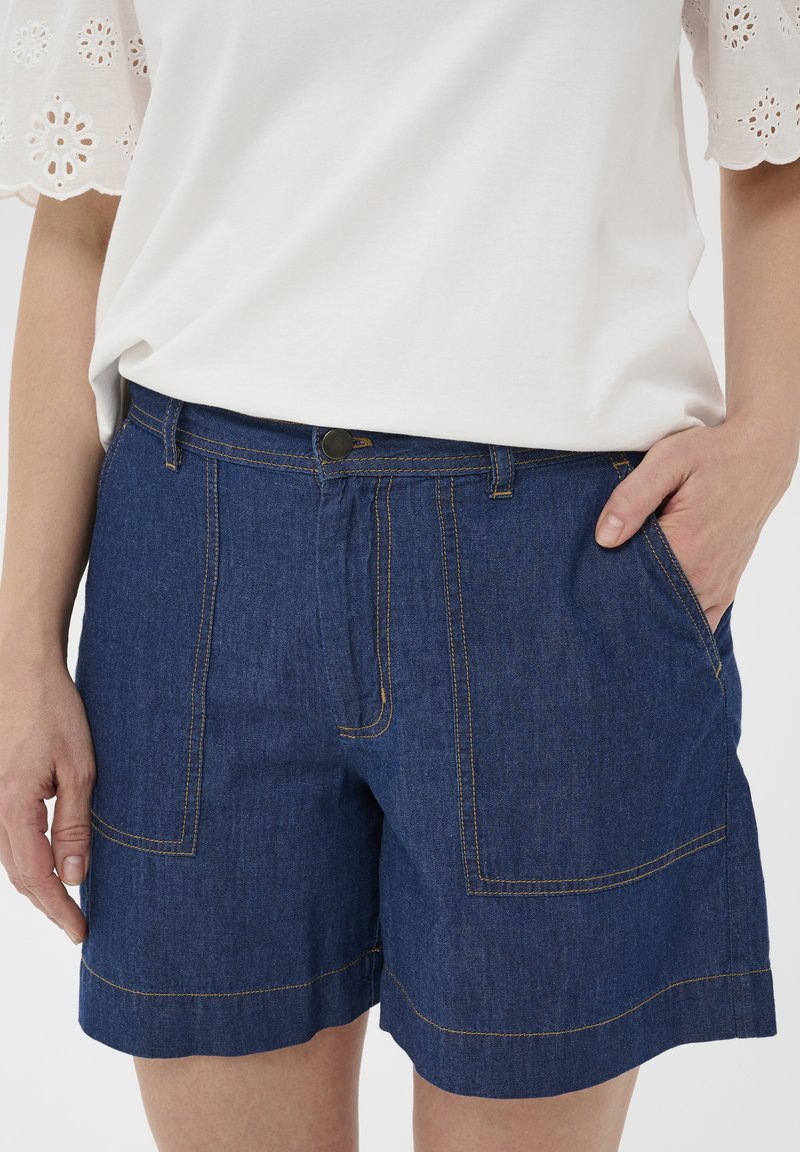 Haut blanc à manches courtes avec détails ajourés, associé à un short en denim bleu doté de grandes poches avant et de surpiqûres visibles.