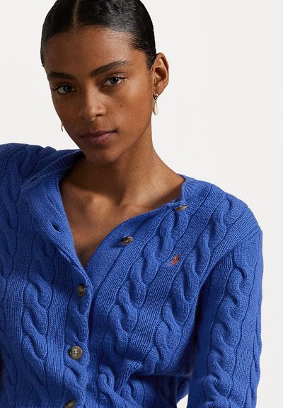 Blauwe gebreide cardigan met een kabelpatroon, uitgevoerd met een sjawl kraag, bruine knopen en een klein rood accentlogo aan de voorkant.