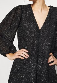 Vestido negro texturizado con escote en V profundo y mangas abullonadas y transparentes, llevado por una persona con las manos en las caderas.